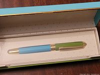 Top view of Kate Spade pen showing light blue barrel, green clip cap with gold accents inside white and gold-trimmed box.