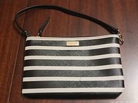 Front view of Kate Spade wristlet showing black and white horizontal stripes, gold-tone logo plaque, and attached black strap on wooden background.