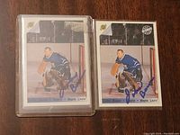 Front view of two Johnny Bower hockey cards in plastic sleeves, showing him in goalie stance in Toronto Maple Leafs uniform, featuring signatures in blue ink.