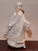 Front and left side view of the white porcelain figurine showing the woman holding a lantern in her left hand, with detailed fur trim on coat and hat.