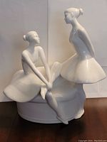Front view of white glazed porcelain ballerina figurine with two ballerinas, one seated and one standing, on an oval base.