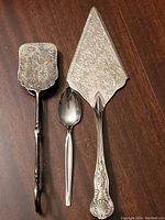 Top side view of the ornate silver cake server, spoon, and salad tongs on a wooden surface.