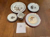 Top view of the 5 anniversary porcelain pieces on a wooden table, showing plates and a cup with floral and anniversary designs, accompanied by a small paper label '21'.