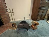 Set of fireplace tools on brass stand, black log holder, and copper bucket on carpeted floor near brick fireplace and wooden cabinet.