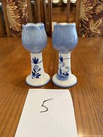 Pair of Delft ceramic candle holders shown from front on wooden table with chairs behind.