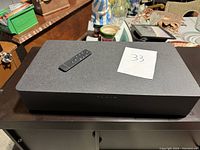 Top and front view of Fluid branded surround sound speaker with remote control on top, showing textured surface and branding.