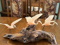 Three pink frosted glass flamingos on a large driftwood base placed on a table with patterned chairs in the background.