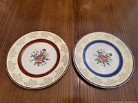 Pair of two English porcelain plates shown side by side on a wooden surface, one with a red band and gold leaf border, the other with a blue band and gold leaf border, both with floral center designs.