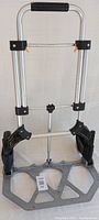 Portable aluminum handcart front view showing foldable handle and base plate with black plastic joints and handle grip.
