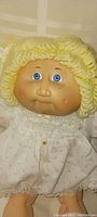 Close-up photo of the doll's head showing yellow yarn hair, blue eyes, molded face, and white dress collar with lace trim.