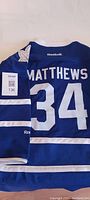 Back view of blue hockey sweater showing 'MATTHEWS' name and large number 34 in white, with Reebok logo on back near collar.