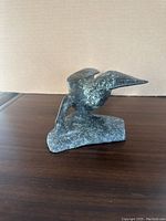 Front angle view of the mottled greenish grey soapstone bird sculpture on a base, showing the bird's head, neck, body, wings, and base.