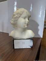 Front view of white marble bust of a girl mounted on a veined black marble base, showing facial details and curls.