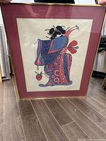 Framed colorful serigraph print depicting a lady in a kimono holding a strawberry, standing on floor with wood plank flooring.