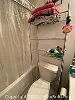 Side view photo showing tall metal tubular frame shelf in bathroom installed over toilet with contents (not included) to illustrate usage.