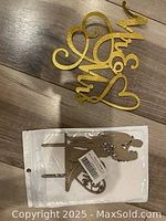 Photo of two unused wedding cake toppers on wooden surface: one gold glitter "Mr & Mrs" topper and one acrylic silhouette bride and groom topper in packaged condition.
