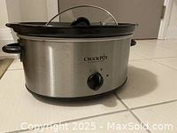 Side view of stainless steel Crock-Pot slow cooker showing black handle, control knob, and glass lid on top.