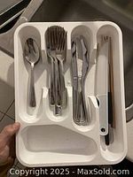 Photo showing stainless steel cutlery including spoons, forks, knives, and a vegetable peeler organized in a white plastic tray.