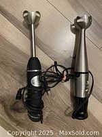 Photo showing two immersion blenders side by side on a wooden floor, highlighting the blending shafts and power cords.