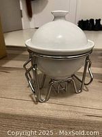 Shows small white ceramic fondue pot with lid on silver metal stand for heating.
