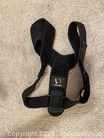 Photo of a black posture corrector brace lying flat on a beige fabric surface. The brace has wide adjustable straps, a padded back support section, and a small label/logo patch.