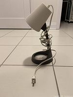 Single small table lamp with grey plastic base and white ribbed lampshade cover. Power cord wrapped around the stand with plug visible.