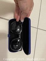 Photo of black oversized round-frame women’s sunglasses held above an open hard protective case with a blue exterior and black interior lining, showing the sunglasses fitting well inside the case.