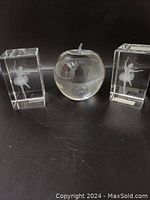 Front view of three crystal paperweights arranged on black surface. Two rectangular blocks with ballerinas flank a clear apple paperweight.