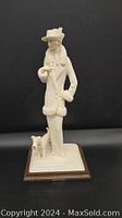 Front view of the white figurine showing woman in long coat and hat standing with terrier dog by her side on wooden base.