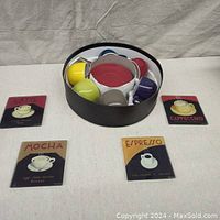 Six colorful espresso cups and saucers in round container with four coffee-themed coasters arranged around it.