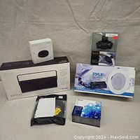 Lot showing boxes of Knocki controller, Edifier MP700 speaker, Pyle ceiling speaker, steering wheel Bluetooth kit, blank DVDs and cable packets on fabric surface