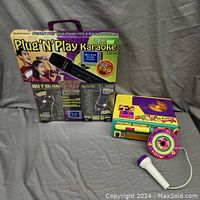 Plug N Play Karaoke system main unit with original box, 4 microphones and instructions, shown with colorful toy karaoke player and microphone on gray fabric background.