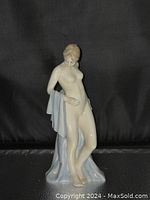 Front view of nude female bone china figurine holding a pale blue draped cloth; standing on a black surface.