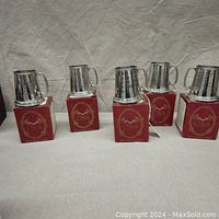 Five pewter child's mugs on top of red boxes, polished finish, visible handles and designs on mugs