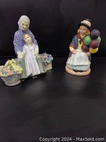 Both figurines side by side, Granny's Heritage with child and flower baskets, Balloon Girl holding balloons