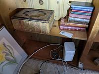 Wide view of wooden shelf containing a large heavily decorated antique Bible and a stack of several vintage hardcover books of various colors and sizes.