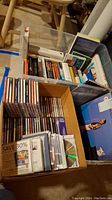 Photo showing assorted hardback books and several CDs packed in a cardboard box, alongside bins containing more books and records.