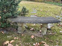 Front view of the curved cement garden bench showing the textured seat and decorative pillar legs with surrounding outdoor ground and leaves.