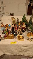 Full view of the Christmas figures including Santa, deer, elves, and bottle brush trees arranged on a white cloth over a table against a basement wall.