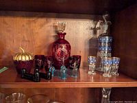 Wide front view showing all colored glass items on a wooden shelf, including red decanter, blue decanter, aperitif glasses, acorn sugar bowl, and utensil rests.