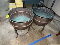 Two copper planters on stands side-by-side showing patina and worn condition.