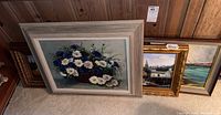 Four framed paintings grouped on floor: one large floral oil painting with a rustic white-gray frame, three smaller gold framed coastal/harbor views.