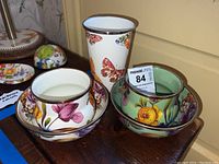 Set of colorful enameled metal cups and bowls arranged on a wooden surface with floral and butterfly prints.