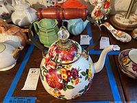 Full view of the floral enameled metal kettle on a table with other teapots visible in background, showing wooden handle and spout, with floral pattern on white base.