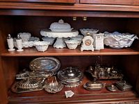 Full shelf display showing the milk glass set including cake pedestal, covered dish, bowls, salt and pepper shakers, pitcher, and vintage Avon bottle mixed with silver items below.