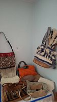 Overview of various handbags and purses piled near a corner wall with a blue and cream woven bag hanging