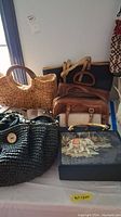 Photo showing a group of handbags of various materials and colors, including woven straw, black cloth, brown leather, and tapestry with elephant design.
