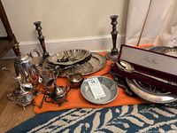 Wide view of all silver plated items including trays, candlesticks, tea set on orange cloth.