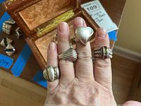 Hand showing four sterling silver rings in different styles including a heart shaped, ridged, and stone-set design.