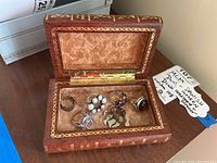 Photo of six rings and one pin displayed inside an open faux book box made of leather-like material with gold detailing and velvet interior.